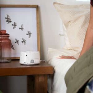 Three-quarter view of Dohm Classic white noise machine showing front dial and vents on bedside table next to bed