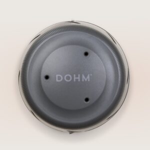 Top view of Dohm charcoal white noise machine showing DOHM logo, three vents, and ridged dial on a light linen background