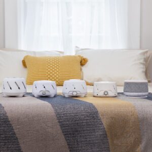Dohm Nova sound machines on a bed showing gray woven fabric covers and glowing night-light rings in a bedroom with pillows