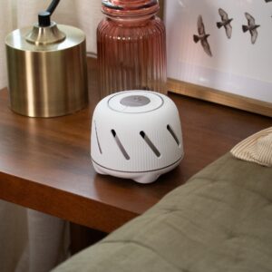 Person using smartphone to control white Dohm Connect sound machine on a wooden bedside table by a bed
