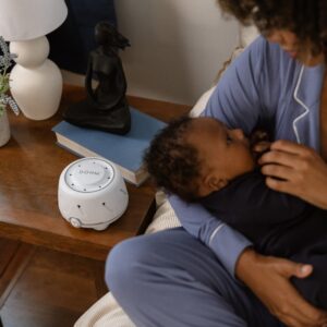 Parent holding a baby beside a Dohm white noise machine on a wooden nightstand in a cozy bedroom