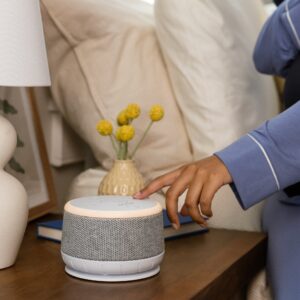 Person reaching for Dohm Nova sound machine and night light on a bedside table with lamp and vase of flowers