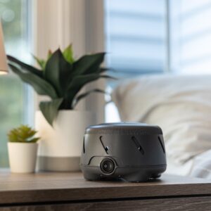 Charcoal Dohm Uno white noise machine on a wooden nightstand beside a lamp and houseplants