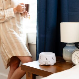 Dohm Uno white noise machine on bedside table next to a lamp and alarm clock, person holding a mug by a window
