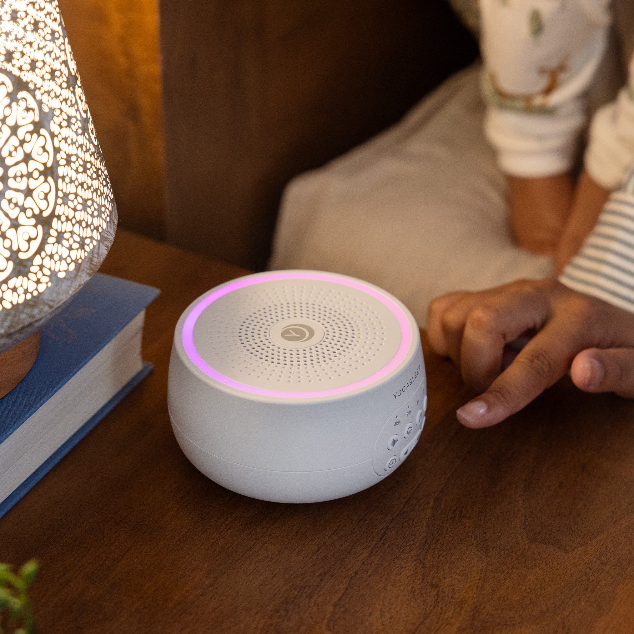 Dreamcenter sound machine glowing purple on a bedside table next to a lamp with a hand reaching to adjust it