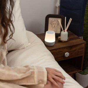 Angled front view of Duet sound machine showing lit warm night light and white top control buttons with gray fabric speaker base on a wooden nightstand beside a bed