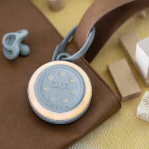 Angled view of Hushh 2 portable baby sound machine showcasing illuminated dimmable nightlight ring and hanging strap on a brown leather bag