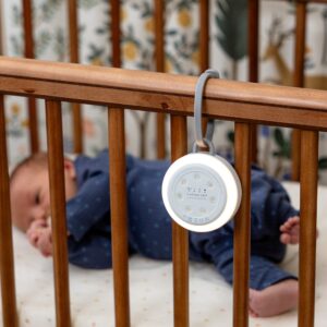 Hushh 2 sound machine hanging from a crib rail by its Yogasleep strap with a baby sleeping in the blurred background