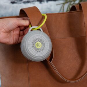 Hushh portable white noise machine clipped to a brown bag strap, gray unit with lime-green power button and loop handle